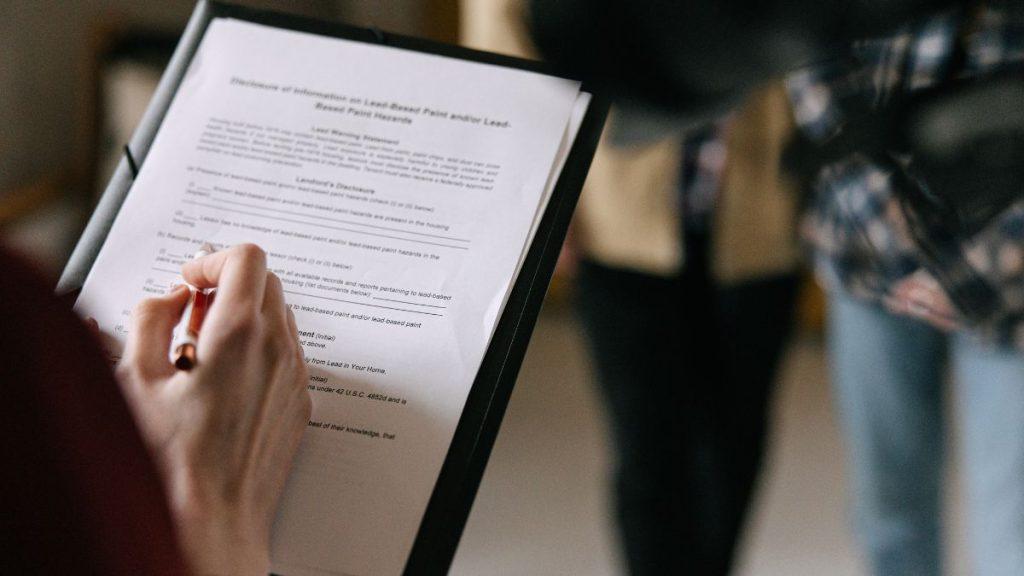 Person writing on a clipboard-held document with a pen in a bright indoor setting