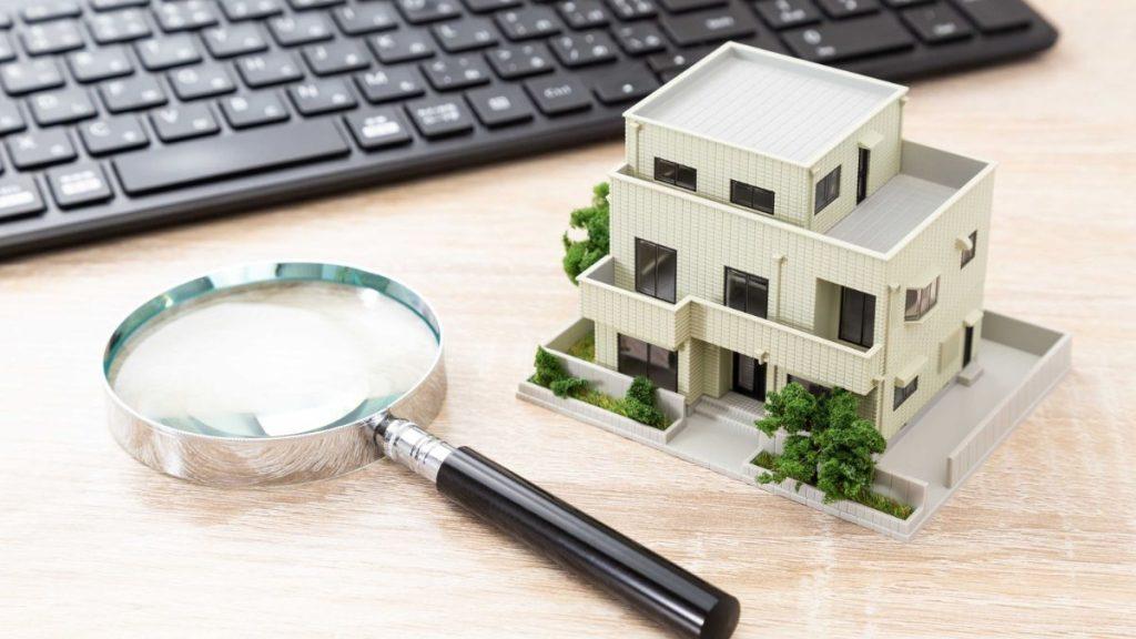 Scale-model house with a magnifying glass on a desk, keyboard in the background, suggesting real estate research or inspection