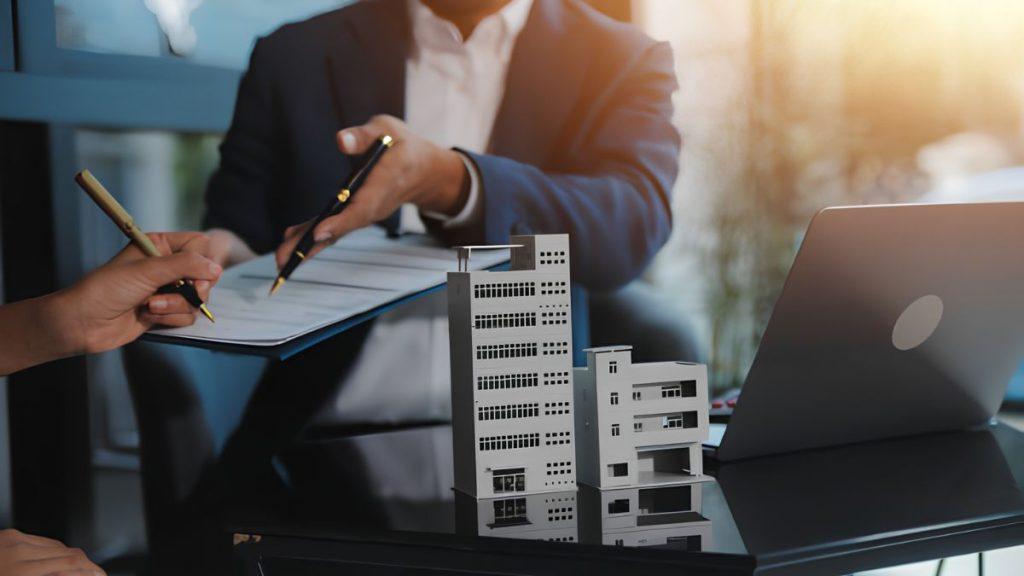 Business meeting with two people drafting documents; miniature buildings model on glass conference table beside a laptop.