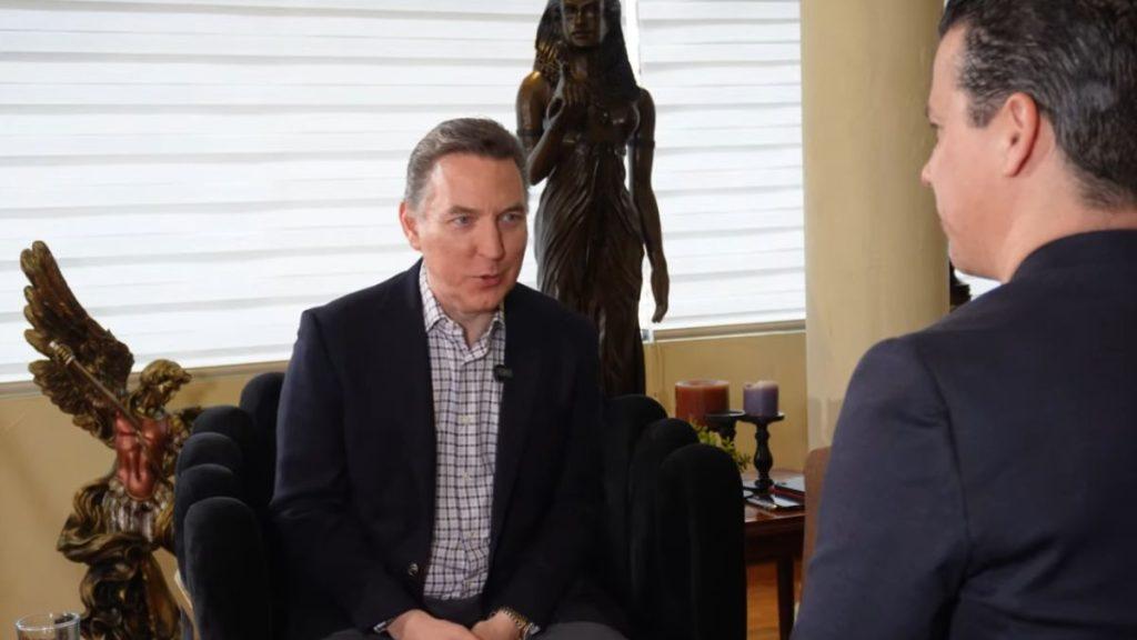Two men in a formal interview scene: one speaks while the other listens, seated in a stylish room with statues and candles in the background.