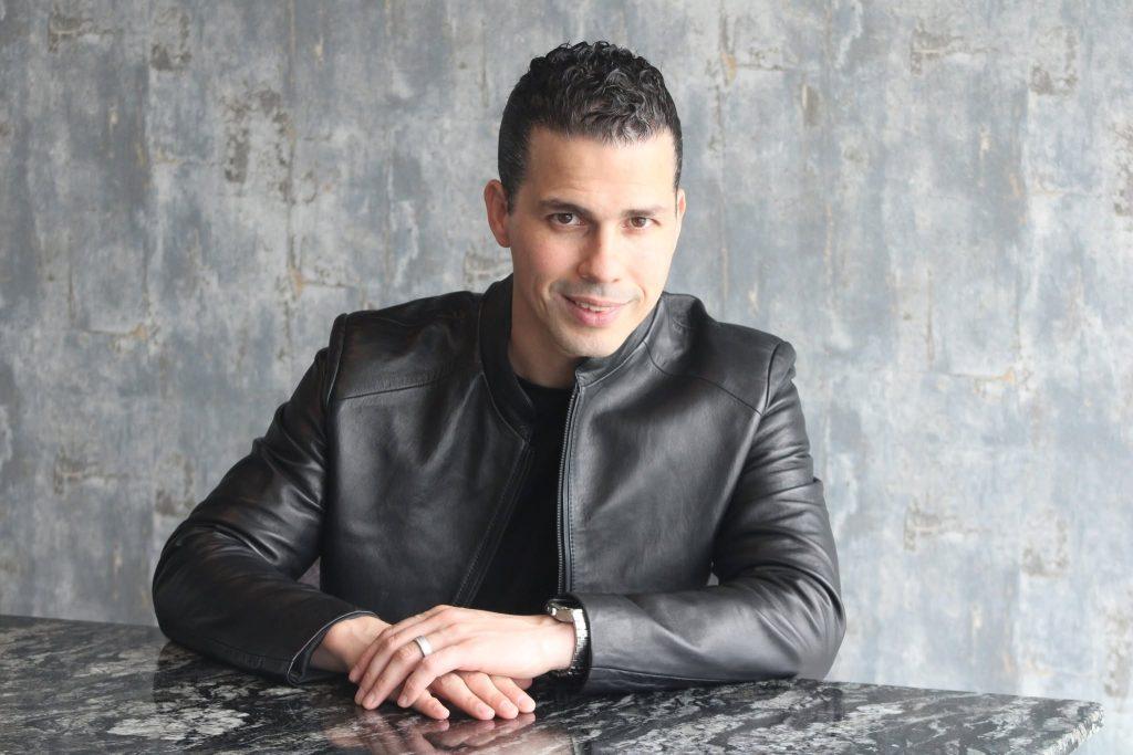 Quantium-Desarrollos-pedro-dávila-vivienda-en-monterrey - El INFORMANTE Man in a black leather jacket sits at a marble table, hands folded, looking at the camera against a grey textured wall.